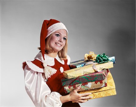1960s YOUNG WOMAN IN RED AND WHITE SANTA HELPER COSTUME AND HAT HOLDING PILE OF WRAPPED CHRISTMAS PRESENTS STUDIO Stock Photo - Rights-Managed, Code: 846-02794273