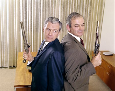 1960s 2 MIDDLE AGED BUSINESS MEN STAND BACK TO BACK HOLDING GUNS PISTOLS DUEL DUELING ANGRY EXPRESSION ENEMY ENEMIES Stock Photo - Rights-Managed, Code: 846-02795169