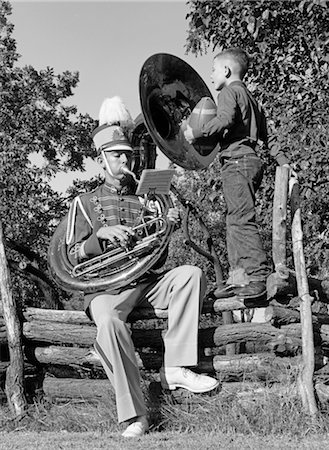 1950s MAN PLAYING A TUBA SITTING ON A LOG FENCE IN A BAND UNIFORM WHILE A BOY WHO IS STANDING ON THE FENCE HOLDING A FOOTBALL WATCHES Stock Photo - Rights-Managed, Code: 846-02795401