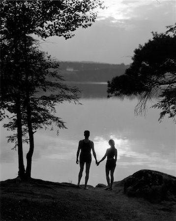 1930s REAR VIEW SILHOUETTE OF MAN AND WOMAN IN BATHING SUITS HOLDING HANDS WATCHING SUNSET LAKESIDE Foto de stock - Con derechos protegidos, Código: 846-02795469