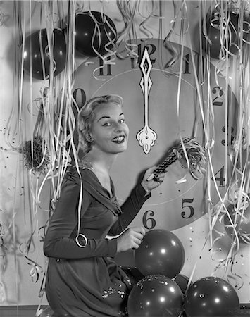 1950s GLAMOROUS WOMAN HOLDING PARTY NOISE MAKER BALLOONS CONFETTI STREAMERS CLOCK AT MIDNIGHT BEHIND HER Stock Photo - Rights-Managed, Code: 846-02795742