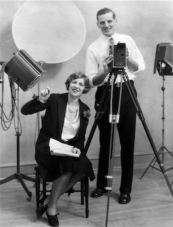 1920s PHOTOGRAPHER STANDING BEHIND CAMERA IN STUDIO WITH ASSISTANT SITTING NEXT TO HIM HOLDING PAPERWORK Stock Photo - Rights-Managed, Code: 846-02795945