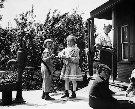 1890s 1900s TURN OF THE CENTURY GROUP OF CHILDREN STANDING ON FRONT PORCH TWO GIRLS PLAYING BY WATER PUMP Stock Photo - Rights-Managed, Code: 846-02795987