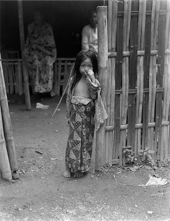 1930s LITTLE JAVANESE GIRL IN BATIK SKIRT STANDING BY BAMBOO FENCE HAND UP TO FACE JAVA INDONESIA Fotografie stock - Rights-Managed, Codice: 846-02796392