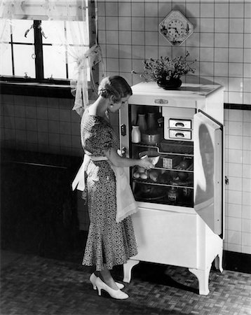 1920s 1930s BLOND WOMAN IN APRON SILK PARTY DRESS HIGH HEELS TAKING DISH FROM ELECTRIC KITCHEN REFRIGERATOR Foto de stock - Con derechos protegidos, Código: 846-02796533