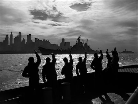 1950s SILHOUETTED SAILORS DECK OF SHIP WAVING SALUTE TO PASSING USN BATTLESHIP AT NIGHT AGAINST NEW YORK CITY SKYLINE Stock Photo - Rights-Managed, Code: 846-02796595