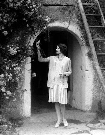 1930s WOMAN IN FLAPPER OUTFIT STANDING IN FRONT OF WHITEWASHED ARCHWAY WITH FLOWERS PICKING BUD BRITTANY FRANCE Stock Photo - Rights-Managed, Code: 846-02796705