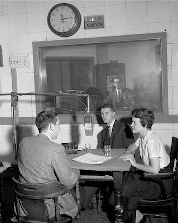 entertainment in the 1960s - ANNÉES 1960 DISC JOCKEY AU MICRO DE RADIO STATION MIKE PARLER AVEC COUPLE HOMME FEMME AT TABLE HOMME DANS LA SALLE DE CONTRÔLE Photographie de stock - Rights-Managed, Code: 846-02796897