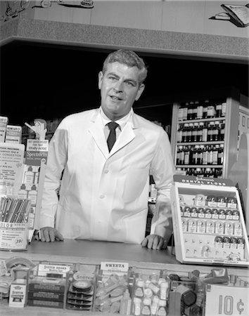 simsearch:846-02796424,k - 1960s PHARMACIST STANDING AT COUNTER FACING CAMERA DRUG COSMETICS SALES CUSTOMER SERVICE LAB COAT PHARMACY STORE RETRO VINTAGE Stock Photo - Rights-Managed, Code: 846-02796917