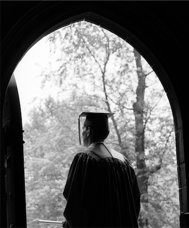 1960ER JAHRE RÜCKANSICHT DES GRADUATE IN DUNKLE GEWÖLBTE TÜR MIT BÄUMEN IM SONNENBESCHIENENEN HINTERGRUND STEHEND Stockbilder - Lizenzpflichtiges, Bildnummer: 846-02797015