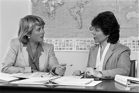 right - ANNÉES 1980 PAIRE D'ENSEIGNANTES AYANT NOTEBOOKS DISCUSSION ÉTALÉE SUR LE BUREAU EN FACE D'EUX CARTE DU MONDE SUR LE MUR EN ARRIÈRE-PLAN Photographie de stock - Rights-Managed, Code: 846-02797165