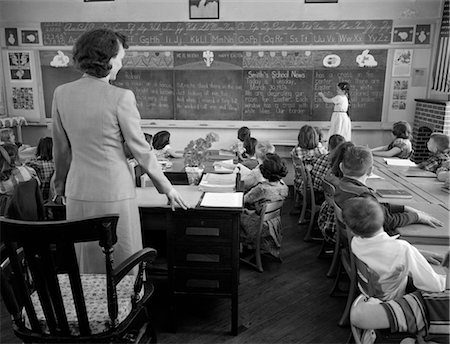 elementary teacher school - 1950s GIRL AT FRONT OF CLASSROOM USING POINTER TO READ THROUGH SCHOOL NEWS ON CHALKBOARD WITH TEACHER STANDING AT DESK Stock Photo - Rights-Managed, Code: 846-02797211