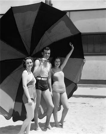 1930s TWO WOMEN ONE MAN SMILING WEARING BATHING SUITS STANDING UNDER EXTRA LARGE STRIPED BEACH UMBRELLA Stock Photo - Rights-Managed, Code: 846-02797291