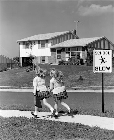 1950s 3 4 BACK VIEW OF TWIN GIRLS IN PLAID SKIRTS & CARDIGANS HOLDING BOOK BAGS RUNNING PAST SCHOOL SLOW SIGN Foto de stock - Con derechos protegidos, Código: 846-02797424