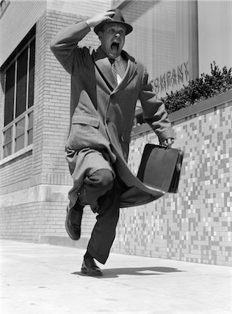 executives running - 1950s FRANTIC MAN RUNNING DOWN STREET HOLDING HAT ON WITH HAND CARRYING BRIEFCASE WEARING TOP COAT COMMUTER BUSINESSMAN Stock Photo - Rights-Managed, Code: 846-02797441