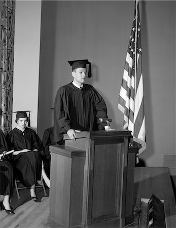 diplôme d'études - ANNÉES 1950 VALEDICTORIAN DONNANT DES DISCOURS AU PODIUM AVEC AUTRES DIPLÔMÉS ASSIS AU FOND DÉTENANT DES DIPLÔMES Photographie de stock - Rights-Managed, Code: 846-02797539