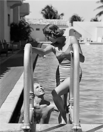 1930s COUPLE WOMAN WEARING ONE PIECE BATHING SUIT CLIMBING LADDER OUT OF SWIMMING POOL LOOKING DOWN AT MAN SMILING MIAMI BEACH FLORIDA Foto de stock - Con derechos protegidos, Código: 846-02797543