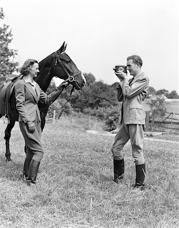 ANNÉES 1940 ÉQUESTRE SOURIANT COUPLE LA FEMME TIENT LA BRIDE DU CHEVAL L'HOMME EST PRISE DE FILMS D'EUX Photographie de stock - Rights-Managed, Code: 846-02797726