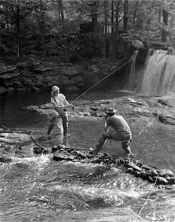 retro fishing - COUPLE MAN TAKING PICTURE PHOTO OF WOMAN FLY FISHING WITH CREEL & WADERS HOLDING ROD IN MIDDLE OF STREAM WITH WATERFALL Foto de stock - Con derechos protegidos, Código: 846-02797721