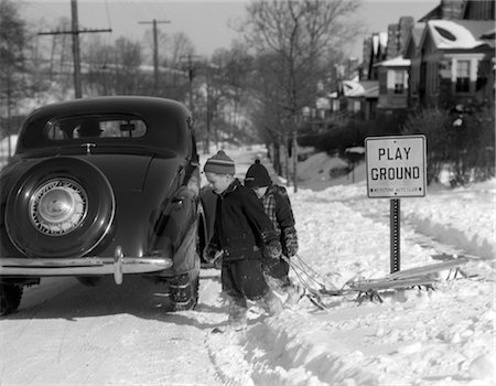 1940s 1950s TWO BOYS PULLING SLEDS ACROSS A SNOWY STREET WITH PARKED CAR AND SIGN THAT SAYS PLAYGROUND SUBURBAN WINTER PLAY Stock Photo - Rights-Managed, Code: 846-02797770