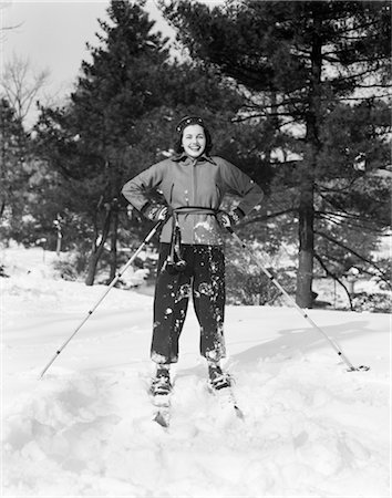 1940s WOMAN ON SKIS HANDS ON HIPS HOLDING SKI POLES ON SKI SLOPE WITH PINE TREES IN BACKGROUND SMILING CRISP FRESH SNOW Stock Photo - Rights-Managed, Code: 846-02797814