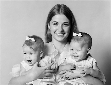 1970s PORTRAIT OF SMILING MOTHER WITH ARMS AROUND TWIN GIRLS WITH BOWS IN HAIR Foto de stock - Con derechos protegidos, Código: 846-02792022