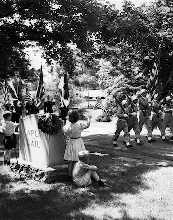 1950s A BOY AND A GIRL STAND BY A TOMBSTONE WAVING TO PASSING COLOR GUARD OF SOLDIERS IN A MEMORIAL DAY PARADE Stock Photo - Rights-Managed, Code: 846-02792285