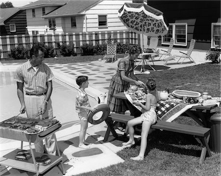 1960s FAMILY OF 4 IN BACKYARD AT POOLSIDE FATHER BARBECUING & MOTHER & CHILDREN MAKING PREPARATIONS AT PICNIC TABLE Foto de stock - Con derechos protegidos, Código: 846-02792288
