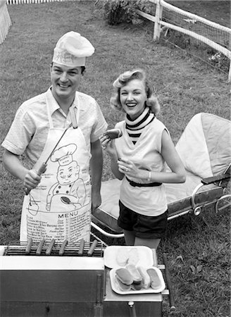 1950s COUPLE BACKYARD GRILL COOK HOT DOGS MAN WEARING APRON TOQUE & SKEWERED HOT DOG Foto de stock - Con derechos protegidos, Código: 846-02792304