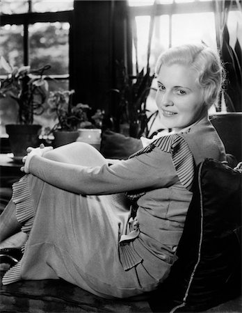 1920s 1930s BLOND WOMAN SMILING SITTING WITH ARMS AROUND HER KNEES IN WINDOW SEAT WITH PILLOWS CUSHIONS & HOUSEPLANTS Foto de stock - Con derechos protegidos, Código: 846-02792976