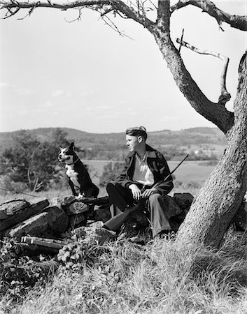 fox-terrier - ANNÉES 1940 GARÇON ASSIS SUR LA CLÔTURE DE PIERRE AVEC LE CHIEN AU REPOS FUSIL ENTRE LES JAMBES DE BOSTON TERRIER Photographie de stock - Rights-Managed, Code: 846-02793002