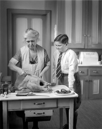 ANNÉES 1930 GRAND-MÈRE EN TABLIER PRÉPARE UNE DINDE DANS LA CUISINE TANDIS QUE LE PETIT-FILS EN ROBE CHEMISE CRAVATE & KNICKERS MONTRES Photographie de stock - Rights-Managed, Code: 846-02793176