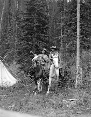 1920s TWO MEN RIDING HORSES THROUGH CAMP GROUNDS IN FOREST COWBOY HAT TENT ADVENTURE COWBOYS BOTH ARE WEARING ANGORA CHAPS Stock Photo - Rights-Managed, Code: 846-02793229