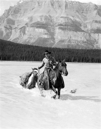 1930s COWBOY WITH BATWING CHAPS ON A BAY HORSE CROSSING A RIVER LEADING A PAINT PACK HORSE WITH MOUNTAINS IN BACKGROUND Foto de stock - Con derechos protegidos, Código: 846-02793220