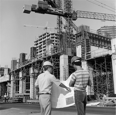 VUE ARRIÈRE DES ANNÉES 1980 DE DEUX TRAVAILLEURS CASQUE DEBOUT DEVANT LE CHANTIER MAJEUR LOOKING OVER BUILDING PLANS D'HOMMES EN PLEIN AIR Photographie de stock - Rights-Managed, Code: 846-02793439