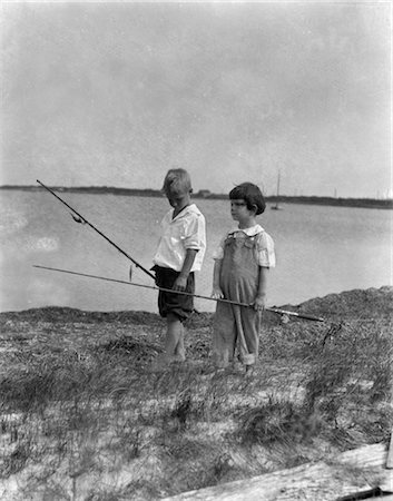 1920s TWO BOYS WALKING ALONG SHORE GOING FISHING POLES BAREFOOT SUMMER FUN FISHING POLES WATER BAY LAKE Fotografie stock - Rights-Managed, Codice: 846-02793607