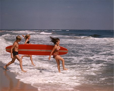 1970s ONE TEENAGE BOY TWO GIRLS IN BATHING SUITS LAUGHING RUNNING INTO OCEAN SURF CARRYING A RED SURFBOARD Foto de stock - Con derechos protegidos, Código: 846-02793865