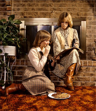 década de 1980 - 1980s 2 TEENAGE GIRLS SISTERS SIT ON HEARTH READING MAGAZINE EAT COOKIES FASHION CLOTHES TEENS BOOTS SKIRTS Foto de stock - Direito Controlado, Número: 846-03164794