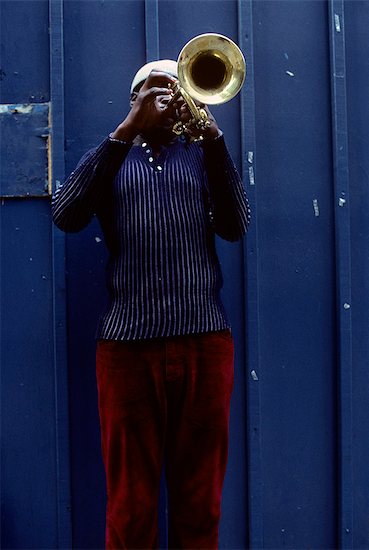 1970s AFRICAN AMERICAN STREET MUSICIAN PLAYING BRASS TRUMPET BY BLUE WALL BLUE SHIRT MAROON PANTS MONOCHROMATIC Stock Photo - Premium Rights-Managed, Artist: ClassicStock, Image code: 846-03165027