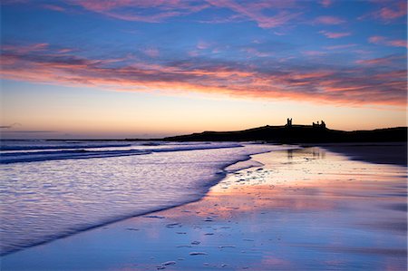 Looking across Embleton Bay at sunrise towards the silhouetted ruins of Dunstanburgh Castle in the distance and the vivid colours in the sky reflecting in the sea and wet sand, Embleton, near Alnwick, Northumberland, England, United Kingdom Photographie de stock - Rights-Managed, Code: 841-07081873