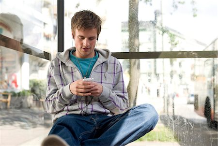 Young man sitting in a bus stop and using a mp3-player, low angle view Stock Photo - Rights-Managed, Code: 853-03459072