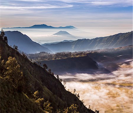 Indonesia, Java, Bromo. A stunning early morning landscape of the Tengger Massif showing the scarp walls of an ancient caldera and volcanoes beyond cloaked in mist.  Low clouds waft over the Sea of Sand in the foreground. Stock Photo - Rights-Managed, Code: 862-07910014