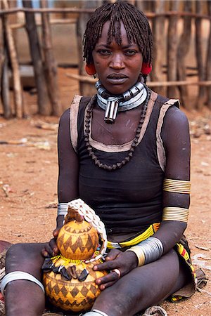 A Hamar woman sits holding a gourd in the village square of Dimeka. Married women wear two heavy steel necklaces. This woman wears an extra necklace with steel a steel phallic symbol which identifies her as a first wife. She wears her hair long in a braided fringe matted with animal fat and ochre. Stock Photo - Rights-Managed, Code: 862-03354047