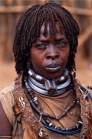 A Hamar woman in the village square of Dimeka. Married women wear two heavy steel necklaces. This woman wears an extra necklace with steel a steel phallic symbol which identifies her as a first wife. She wears her hair long in a braided fringe matted with animal fat and ochre. Foto de stock - Con derechos protegidos, Código: 862-03354048