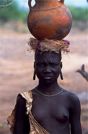 Une jeune fille de la tribu Mursi. Cette fille est célibataire car elle n'a pas la lèvre inférieure percée et plaque de lèvre d'argile d'une femme mariée. Dans la vallée de l'Omo, les Mursi ont la réputation d'être extrêmement violent et agressif, mais aussi pour être qualifiés dans la fabrication de pots en argile. Cette fille porte un pot sur la tête à l'aide d'un anneau d'herbes tressées pour aider à prévenir le pot de tomber. Photographie de stock - Rights-Managed, Code: 862-03354059