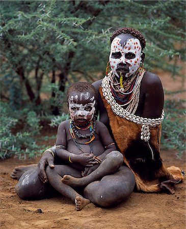 A Karo woman sits with child. A small Omotic tribe related to the Hamar,who live along the banks of the Omo River in southwestern Ethiopia,the Karo are renowned for their elaborate body painting using white chalk,crushed rock and other natural pigments. Typically for a Karo woman,the mother has ochred her hair in tight ringlets and has a ring through her bottom lip. Foto de stock - Direito Controlado, Número: 862-03354094