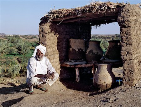 A Nubian man squats beside earthenware water containers,which keep water cool in the heat of the day. The village where he lives on the River Nile,close to Shaallal arraabia or the 4th Cataract,will be flooded in 2008 when a huge Arab funded,Chinese-built,hydroelectric dam will be completed. Already,some villages have been relocated to irrigated land far away. Stock Photo - Rights-Managed, Code: 862-03354586