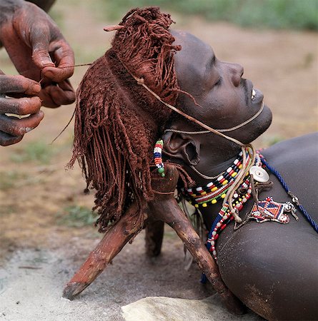 A Samburu warrior has his Ochred hair braided by a friend. A mixture of cow's urine and ashes is often rubbed into the hair first to help straighten it. The wooden headrest is used as a pillow at night. Long braids of Ochred hair distinguish warriors from other members of their society. The warriors are vain and proud,taking great trouble over their appearance. Stock Photo - Rights-Managed, Code: 862-03366042