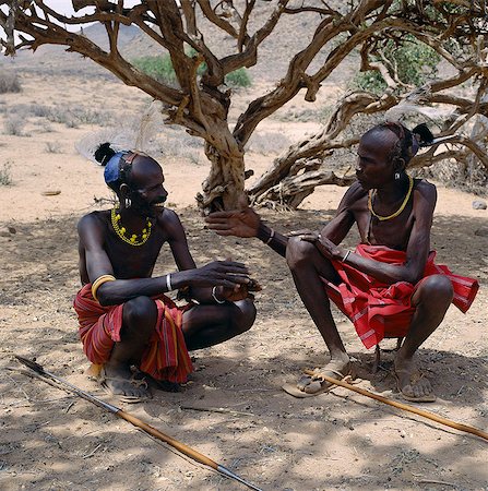 Two Turkana men in traditional attire relax in the heat of the day under a shady tree. Every man will have a wooden stool,which doubles up as a pillow at night to protect his clay hairdo. Men will never sit on the ground; only women and children are permitted to do so. Stock Photo - Rights-Managed, Code: 862-03366103