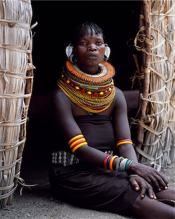 A Turkana woman,typically wearing many layers of bead necklaces and a series of hooped earrings with an pair of leaf-shaped earrrings at the front,sits in the entrance to her hut. Foto de stock - Con derechos protegidos, Código: 862-03366494